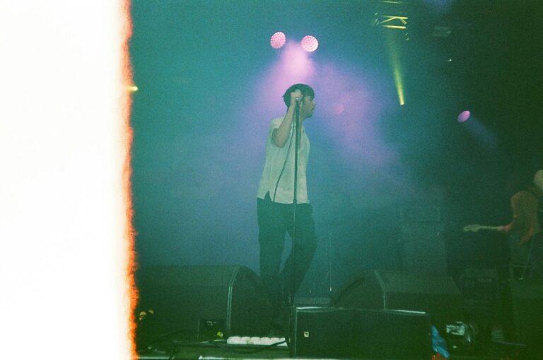 A musician energetically performs on stage with colorful lights and smoke, filmed in an analog style.